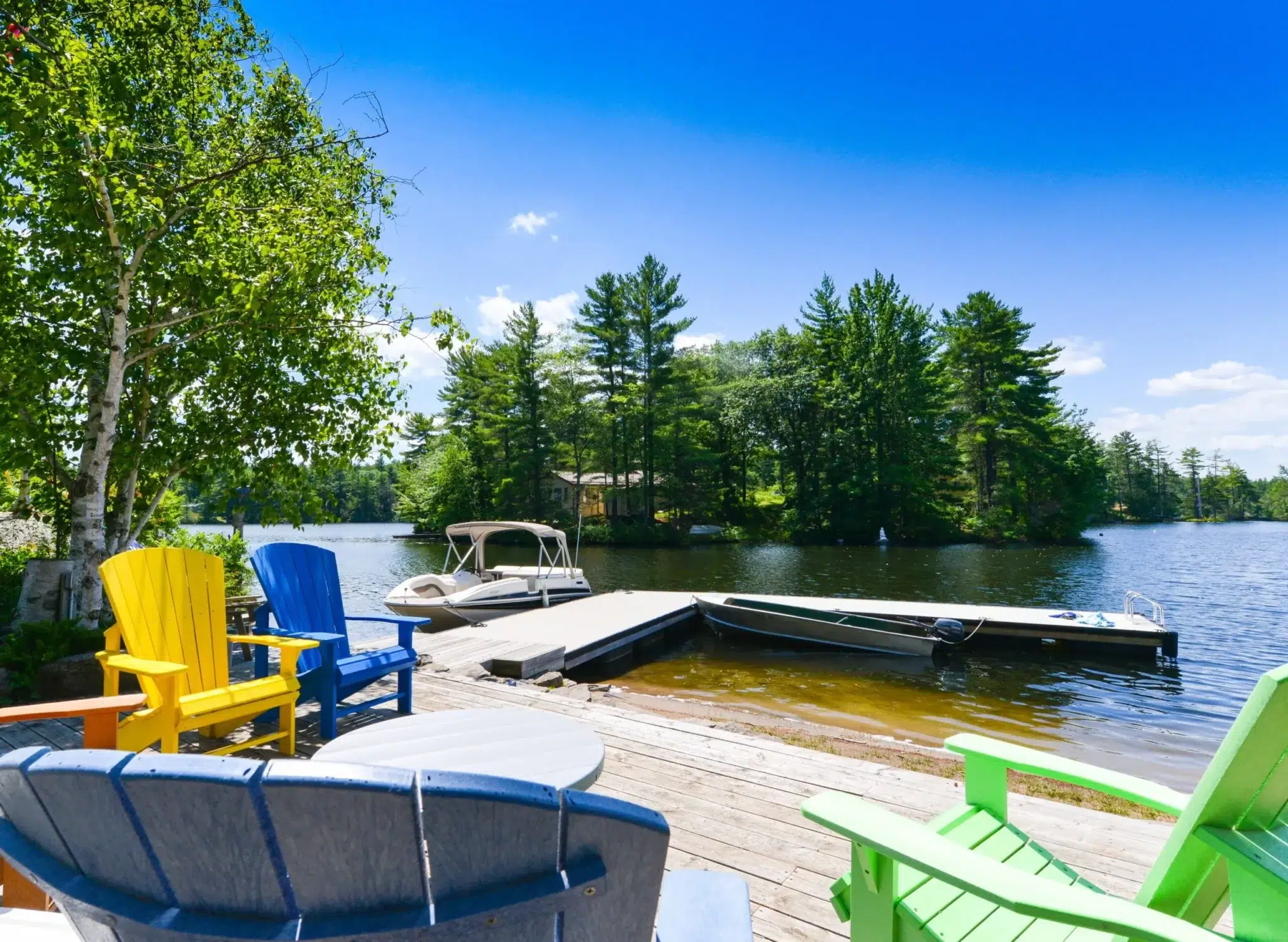 A colorful outdoor seating area on an aluminum deck, overlooking a lake with a dock and a boat under a bright blue sky.