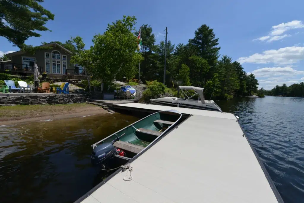 A view of a lakeside cottage with a pontoon boat and a small rowboat tied to a dock made of Aluminum Decking.
